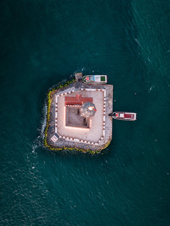 Aerial shot of a lighthouse on an island with boats docked nearby in the ocean.